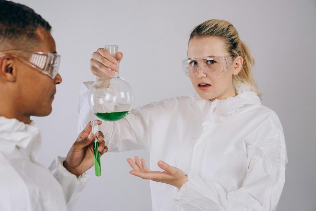 Two scientists in protective gear conduct an experiment with green liquid in a lab setting.